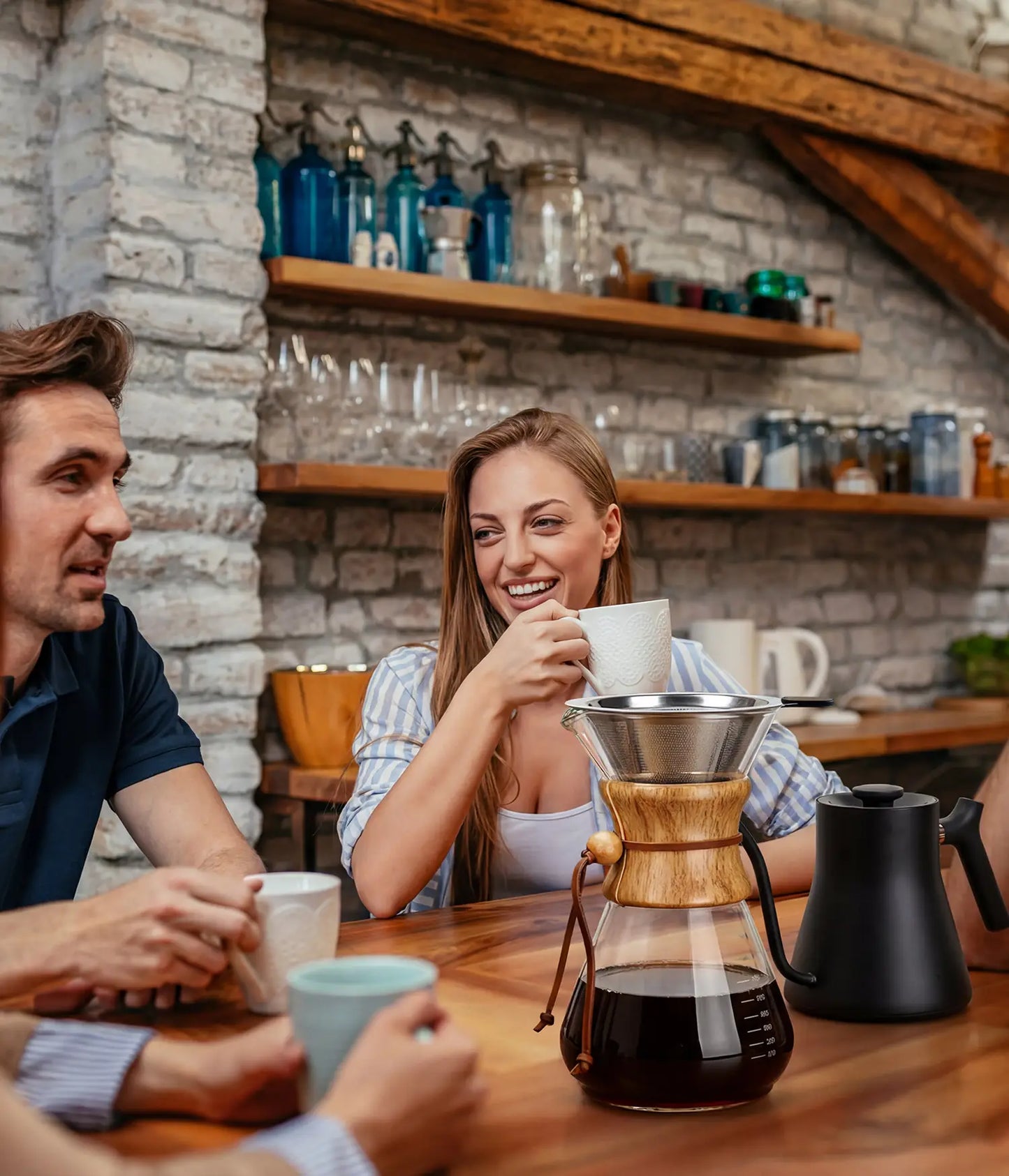 Friends laughing and sharing a generous brew around a rustic wooden table, featuring the elegant AGOGO 1000ml slim glass pour over carafe.