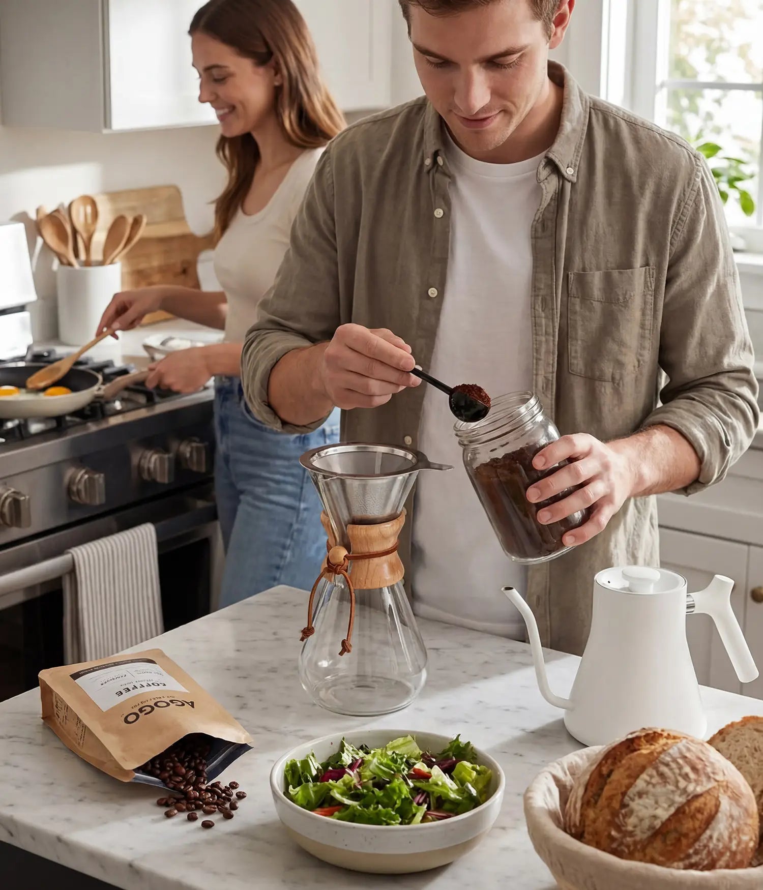Couple brewing morning coffee in an earth-tone home kitchen, adding fresh coffee grounds from an AGOGO bag into the 600ml glass pour over dripper.