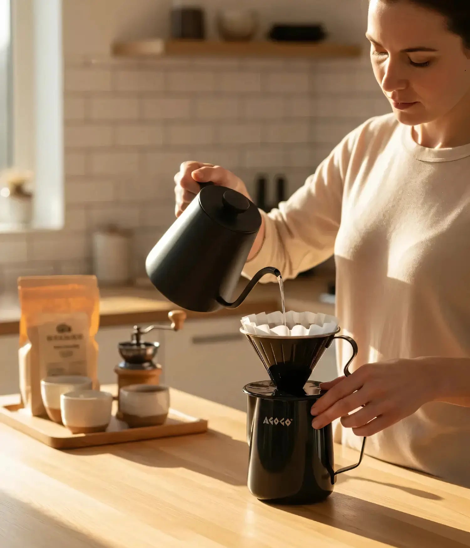 Woman brewing a clean cup of coffee with the unbreakable AGOGO matte black stainless steel pour over set and paper filter in a sunlit, earth-tone kitchen.