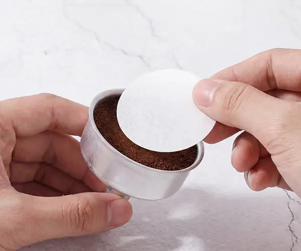 Close-up of hands placing a round AGOGO precision paper filter on top of the coffee grounds in the moka pot funnel for a cleaner extraction.