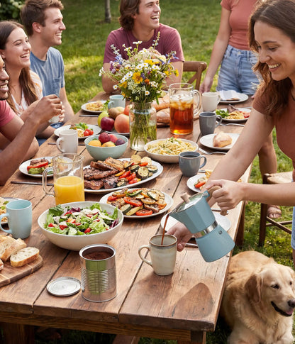 A woman pouring coffee from the lake blue AGOGO Milano Moka Pot during a joyful backyard garden party with friends, a large feast on a wooden table, and a golden retriever.