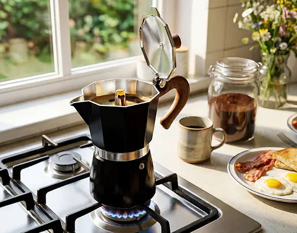A matte black AGOGO Milano Moka Pot with an open lid, brewing rich espresso on a gas stove next to a morning breakfast spread.