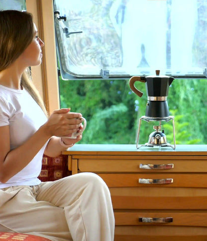 A woman relaxing in an RV looking out the window with a mug of coffee, while the matte black AGOGO Moka Pot rests on a small portable burner nearby.