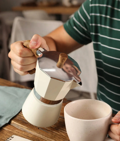 Close-up of a person pouring freshly brewed espresso from the warm cream AGOGO Milano Moka Pot into a ceramic mug on a wooden rustic table.