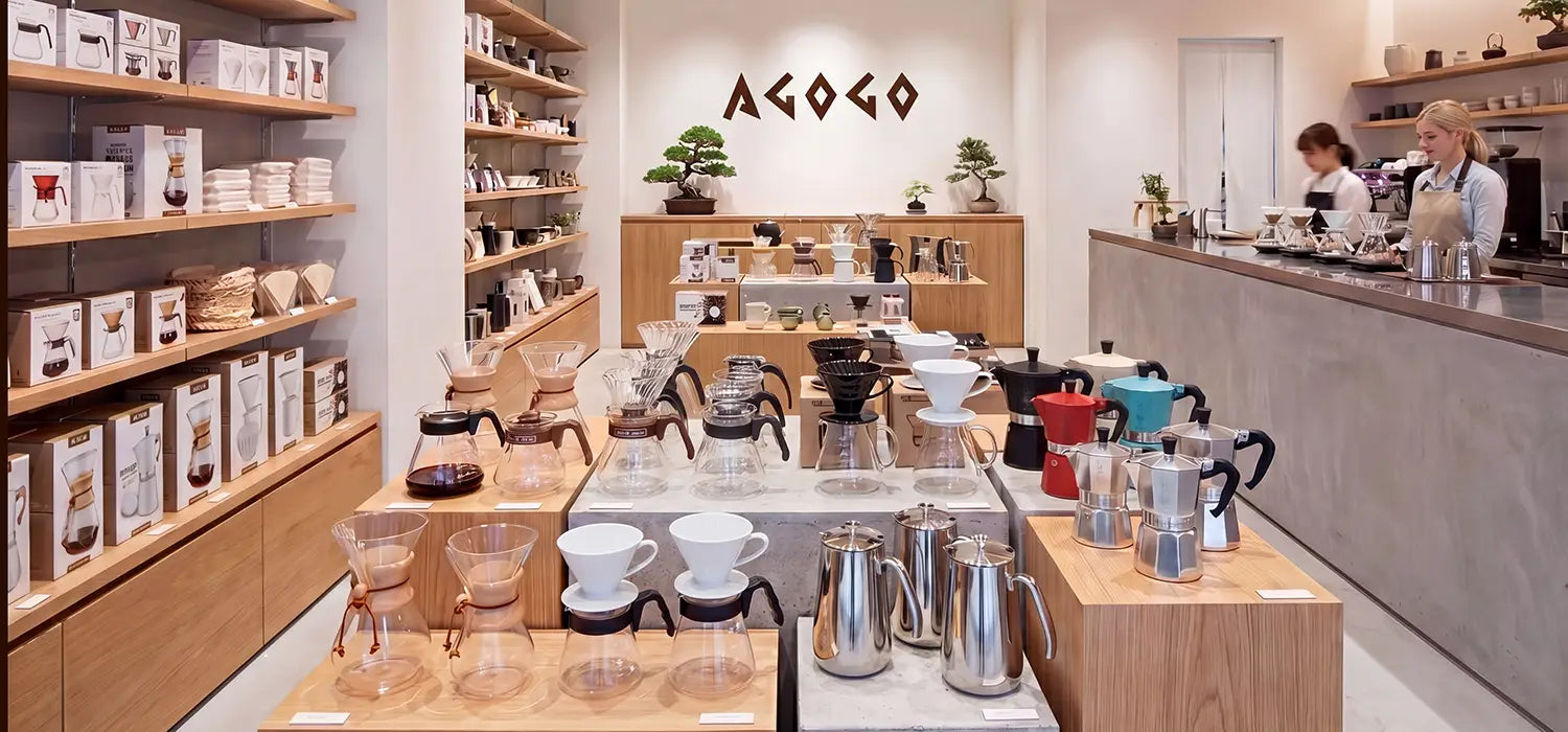 A bright, modern specialty cafe and retail boutique beautifully displaying AGOGO premium coffee gear, including classic moka pots, glass pour-over drippers, and stainless steel servers on wooden shelves.