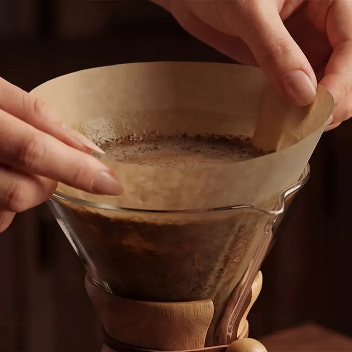 Two hands gently lifting a wet circle coffee filter from a glass brewer to break the vacuum seal and restore steady flow.