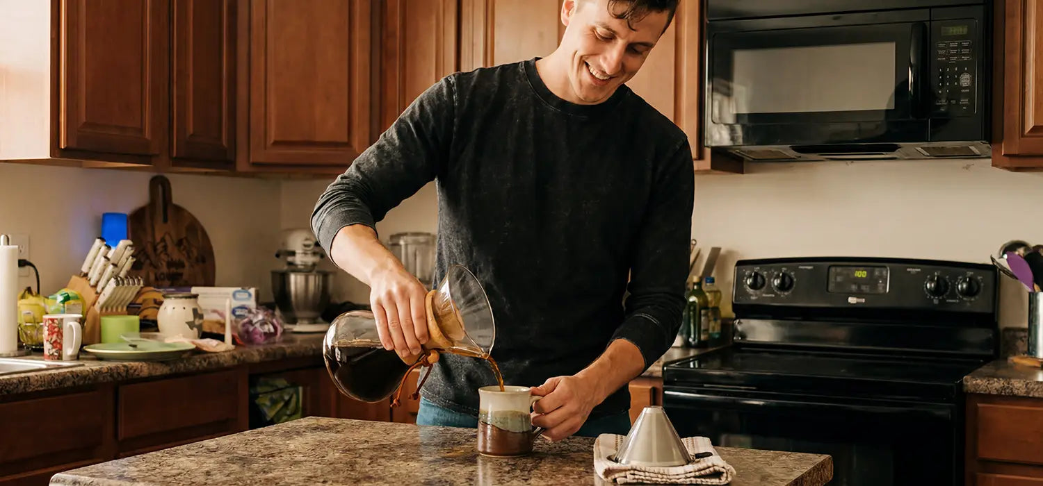 A smiling man in a warm home kitchen pours freshly brewed coffee from an AGOGO glass pour-over server into a ceramic mug, with a stainless steel filter resting on the counter.