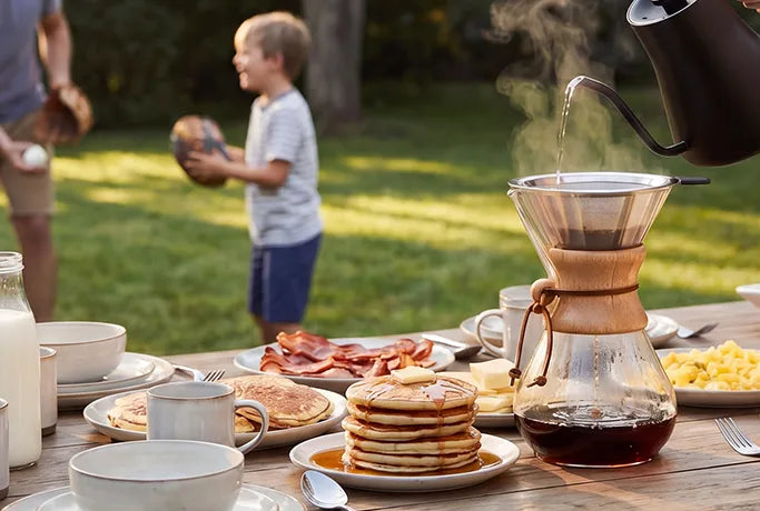 Pouring hot water into an AGOGO stainless steel coffee filter over a glass carafe during a sunny outdoor family breakfast.