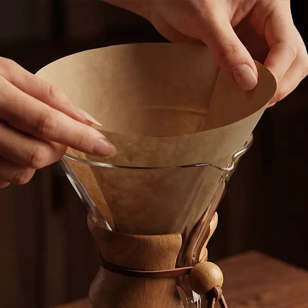 Hands gently placing a natural brown, pre-folded half-moon circle coffee filter seamlessly into a classic glass pour-over brewer.